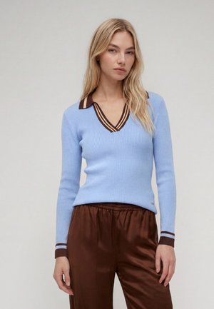 Femme blonde portant un pull côtelé bleu clair avec un col et des poignets rayés marron, associé à un pantalon en satin marron sur un fond uni.