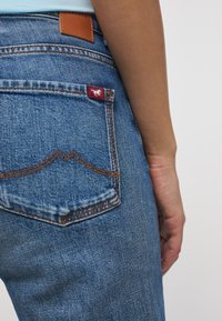 Niebieskie dżinsy z tylną kieszenią, która ma pomarańczowe szwy, czerwoną metkę z białym logo oraz brązową skórzaną etykietę na pasku.