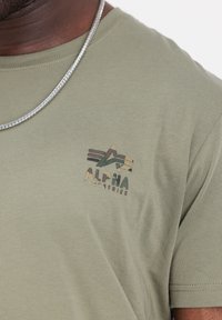 Olivgrön bomulls-t-shirt med en flerfärgad "ALPHA INDUSTRIES"-logotyp. Kort ärm och klassisk rund halsringning. Silverkedja synlig.