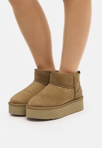 Bottines en daim marron à bout rond, tige courte, et semelle compensée texturée, avec un design simple à coutures autour de la partie supérieure.