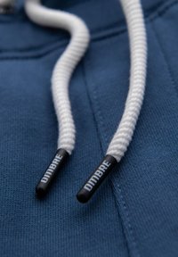 Blauwe hoodie met witte katoenen trekkoorden met zwarte plastic punten, waarop het woord "DIMBRE" is gedrukt. De textuur is glad en gebreid.