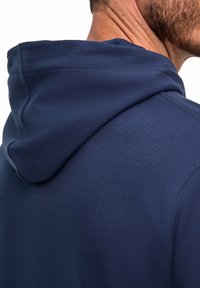 Primo piano della spalla e del collo di un uomo che indossa un indumento con cappuccio di un blu scuro texture, mostrando i dettagli del tessuto e delle cuciture.