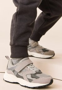 Szare sportowe buty z siateczkowym i zamszowym wierzchem, z rzepem, białą podeszwą środkową i teksturowaną czarną gumową podeszwą.