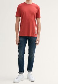 Červené bavlněné tričko s krátkým rukávem, s bočním grafickým potiskem v bílé barvě, kombinované s modrými džínami a bílými teniskami.