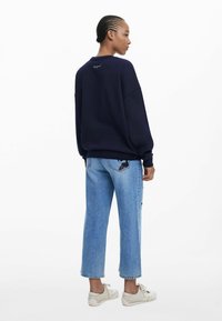 Marineblaues Sweatshirt mit geripptem Kragen, lässiger Passform und gesticktem Detail auf der Rückseite; hellblaue Jeans mit Patches; beigefarbene Sneakers.