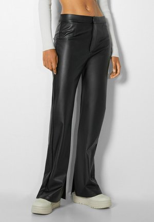 Frau trägt schwarze Lederhose mit weitem Bein und vorderen Schlitzen, dazu weiße Plateau-Sneaker, steht vor hellem, einfarbigem Hintergrund.