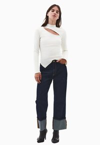 Witte lange mouwen top met een diagonaal uitgesneden ontwerp, gecombineerd met donkerblauwe wijde jeans met opgerolde denimdetails. Zwarte haklaarzen.