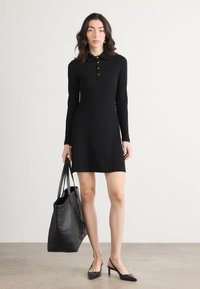 Zwarte polokleed met lange mouwen en een rechte silhouette, met gouden knopen bij de kraag, gecombineerd met een grote zwarte handtas en zwarte hakken.