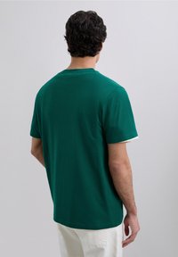 Man met donker krullend haar, gekleed in een groene korte mouw shirt en witte broek, staande met het gezicht naar een effen lichtgrijze achtergrond.