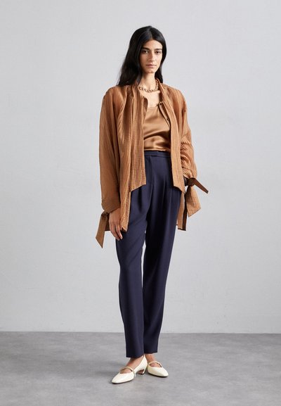 Brown textured, open-front shirt over a gold satin top, paired with navy trousers and white shoes with a gold accent. Simple chain necklace visible.