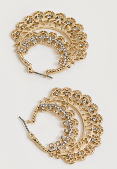 Boucles d’oreilles créoles en or avec des motifs filigranés complexes et des strass clairs incrustés sur un fond clair.