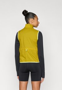 Κίτρινο μεσαίο στρώμα γιλέκο με ανακλαστικές λεπτομέρειες, κλείσιμο με φερμουάρ και ελαστικό κορδόνι στη μέση, σε συνδυασμό με μαύρα μακρυμάνικα και σορτς.