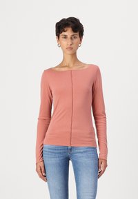Haut à manches longues de couleur rose pâle en tissu doux, avec un décolleté large et un détail de couture à l'avant, associé à un jean bleu clair.