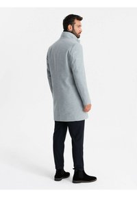 Cappotto in lana grigia con colletto alto, vestibilità slim e chiusura a singolo bottone. Abbinato a pantaloni scuri e stivali neri alla caviglia.