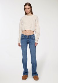 Kremowy sweter w warkocze, niebieskie dżinsy dzwony i brązowe zamszowe buty. Ubranie ma teksturowane materiały i luźny krój.