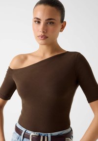 Brązowy prążkowany top z odkrytymi ramionami i krótkimi rękawami, o dopasowanym fasonie. Połączony z jasnoniebieskim dżinsem i ciemnym paskiem.