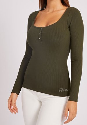 Femme portant un top moulant côtelé vert olive à manches longues avec un col rond, trois boutons et un pantalon blanc ajusté.