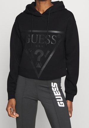 Personne portant un sweat à capuche Guess noir avec un grand logo et des leggings gris foncé avec l'inscription Guess blanche sur la cuisse.