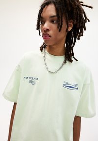 Lys grøn bomulds t-shirt med korte ærmer, prydet med blå grafisk tekst og design foran, båret med en sølv kædeste.