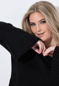 Schwarzer gerippter Pullover mit weiten Ärmeln und einem strukturierten unteren Saum. Das Modell hält die Hände nah am Ausschnitt. Weiches Strickmaterial.