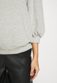 Sweat-shirt gris en mélange de coton avec manches bouffantes et poignets côtelés, associé à un pantalon en cuir lisse noir ; coutures détaillées visibles.