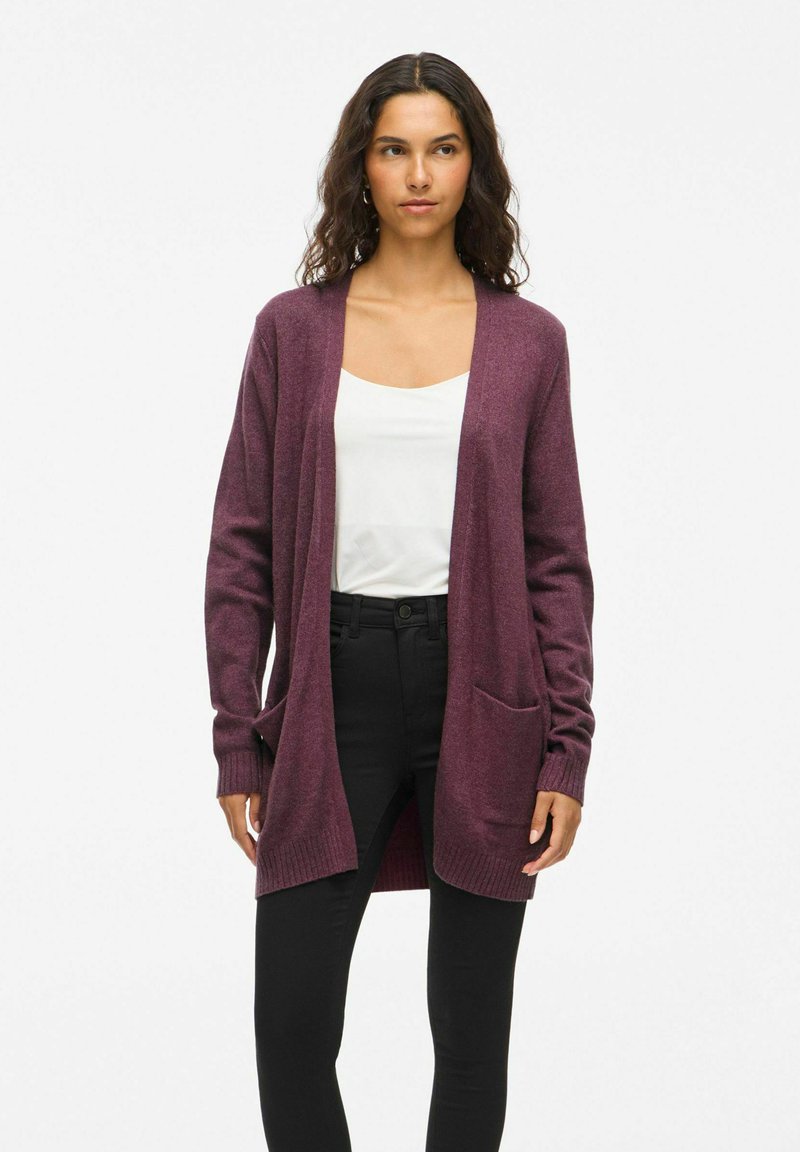 Cardigan en maille bordeaux à manches longues, à ouverture devant, avec deux poches avant ; texture douce avec des poignets côtelés. Porté par-dessus un haut blanc et un pantalon noir.