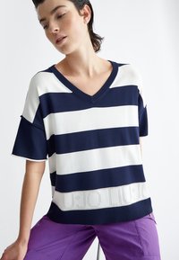 Maglietta a righe blu navy e bianco in maglia con scollatura a V, maniche corte e orlo texturizzato con lettere ritagliate. Abbinata a pantaloni viola.