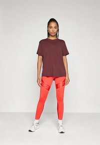 Bordowa koszulka z krótkim rękawem w zestawieniu z jasnoczerwonymi legginsami z odblaskowym logo. Modelka nosi białe buty sportowe.