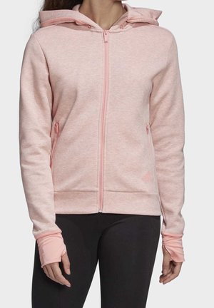 Frau trägt ein hellrosa Kapuzen-Sweatshirt mit Reißverschluss und Daumenlöchern sowie schwarze Leggings, steht vor einem schlichten hellen Hintergrund.