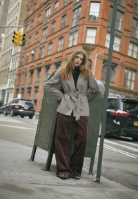 Kariertes Blazer mit Gürtel, dunkler Rollkragenpullover, weite bordeauxfarbene Cordhosen und glänzende schwarze Schuhe. Stadtlandschaft mit roten Backsteingebäuden.