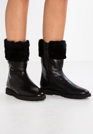 Czarne, skórzane kozaki sięgające do połowy łydki z grubymi gumowymi podeszwami i czarnymi mankietami z futra przewiniętymi do góry, noszone przez osobę stojącą na jasnej powierzchni.