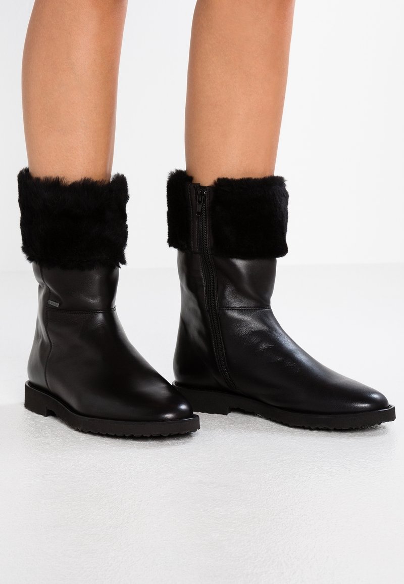 Bottes en cuir noires mi-mollet avec semelles en caoutchouc épaisses et revers en fourrure noire, portées par une personne debout sur une surface claire.