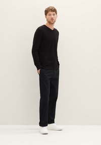 Zwarte V-hals trui, slim-fit zwarte broek en witte sneakers. Model staat met de handen in de zakken tegen een neutrale achtergrond.