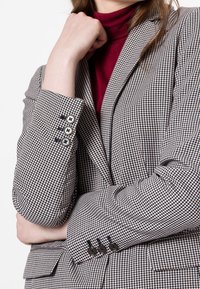 Blazer en patrón de ajedrez blanco y negro, con solapas muesca, puños de tres botones y suave textura de tela.
