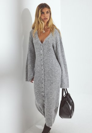 Gray ribbed knit cardigan dress with a V-neck, full-button front, and wide sleeves. Model holds a black tote bag, standing against a white background.