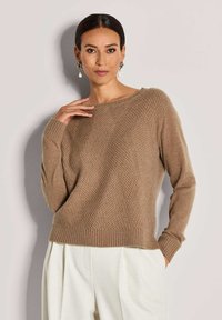 Eine Frau trägt einen strukturierten braunen Strickpullover und weiße Hosen. Sie posiert mit einer Hand an ihrem Hals und der anderen in ihrer Tasche.