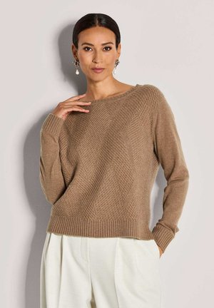 Eine Frau trägt einen strukturierten braunen Strickpullover und weiße Hosen. Sie posiert mit einer Hand an ihrem Hals und der anderen in ihrer Tasche.