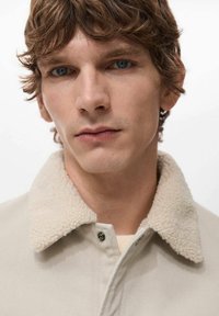 Beige Jacke mit texturiertem, cremefarbenem Lammfellkragen; sichtbarer Knopfverschluss; Modell hat hellbraunes, welliges Haar und einen gleichmäßigen Hautton.