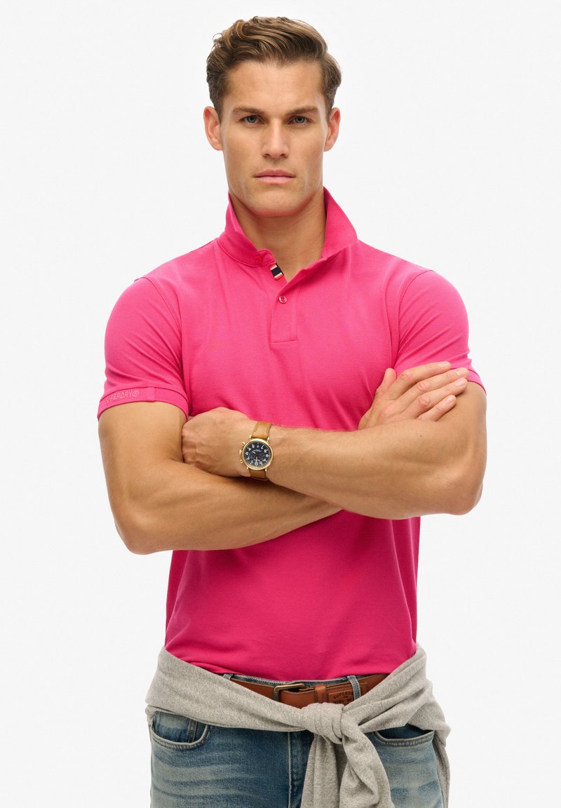 Lyserød kortærmet polo t-shirt med krave, der har et mærke på ærmet. Kombineret med en grå sweater, der er bundet om livet.