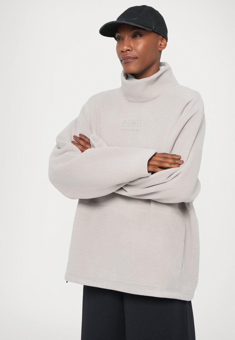Personne portant un sweat à col montant gris clair et une casquette noire, se tenant les bras croisés devant un fond blanc uni.