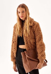 Bruine, pluizige jas met een gestructureerde afwerking, gecombineerd met een bruine leren tas. Een zwarte cropped top en hoge zwarte broeken maken de look compleet.