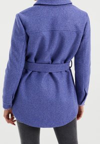 Fialový vlněný kabát s páskem, má klopový límec, dlouhé rukávy a mírně zaoblený lem. Texturovaná tkanina s jemným povrchem.