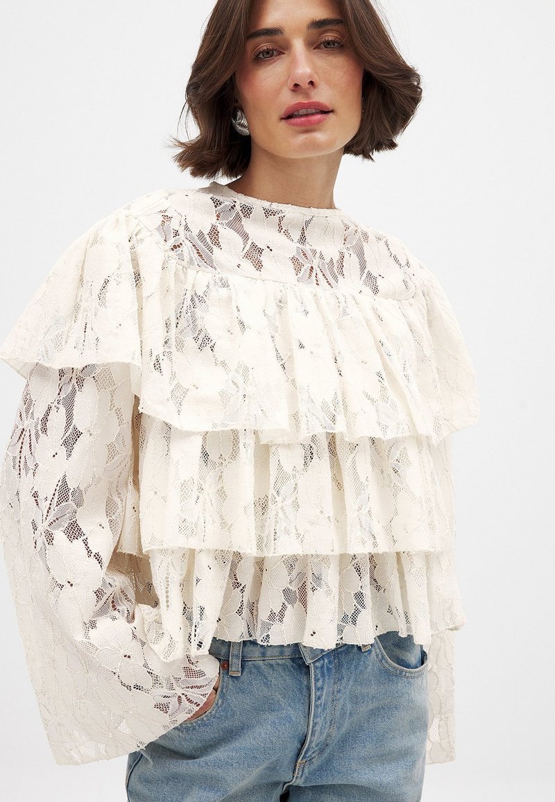 Blouse en dentelle blanche avec trois volants superposés, manches longues et col montant. Assortie à un jean en denim bleu clair.