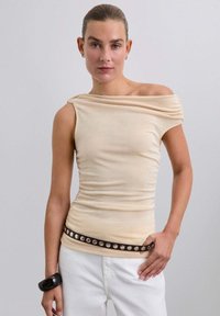 Top beige sin mangas con escote drapeado, diseño ajustado, textura fruncida, combinado con un cinturón marrón con acentos metálicos redondos.