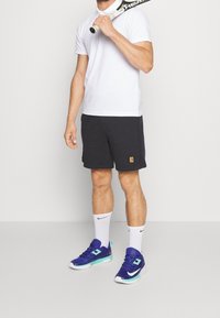 T-shirt polo blanc, shorts noirs avec logo, chaussettes blanches et chaussures de sport bleues avec accents turquoise. Tenant une raquette de tennis.