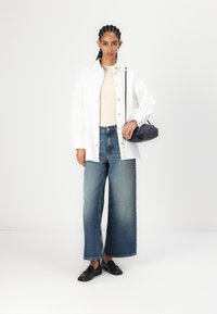 Cămașă albă supradimensionată cu nasturi, bluză bej, blugi largi albaștri și mocasini negri. O geantă mică neagră tip crossbody completează ținuta.