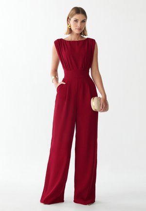 Femme portant une combinaison rouge sans manches, des boucles d’oreilles et des bracelets dorés, tenant une petite pochette ronde, debout devant un fond uni.