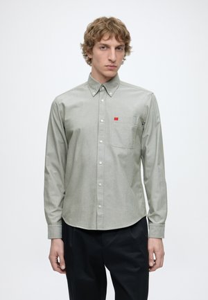 Jeune homme aux cheveux bouclés portant une chemise gris clair boutonnée avec une poche poitrine et un pantalon noir, debout devant un fond blanc uni.