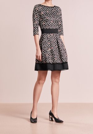 Robe noire avec un motif floral blanc, manches trois-quarts, taille cintrée, jupe évasée, ceinture noire et ourlet noir. Portée avec des chaussures à talons noires.