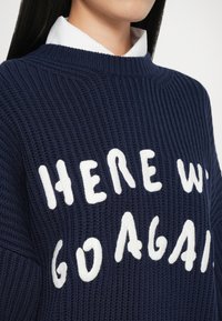 Marine strikket sweater med hvid tekst "HER GÅR VI IGEN." Nærbillede, der fremhæver ribstrukturen og den runde halsudskæring.